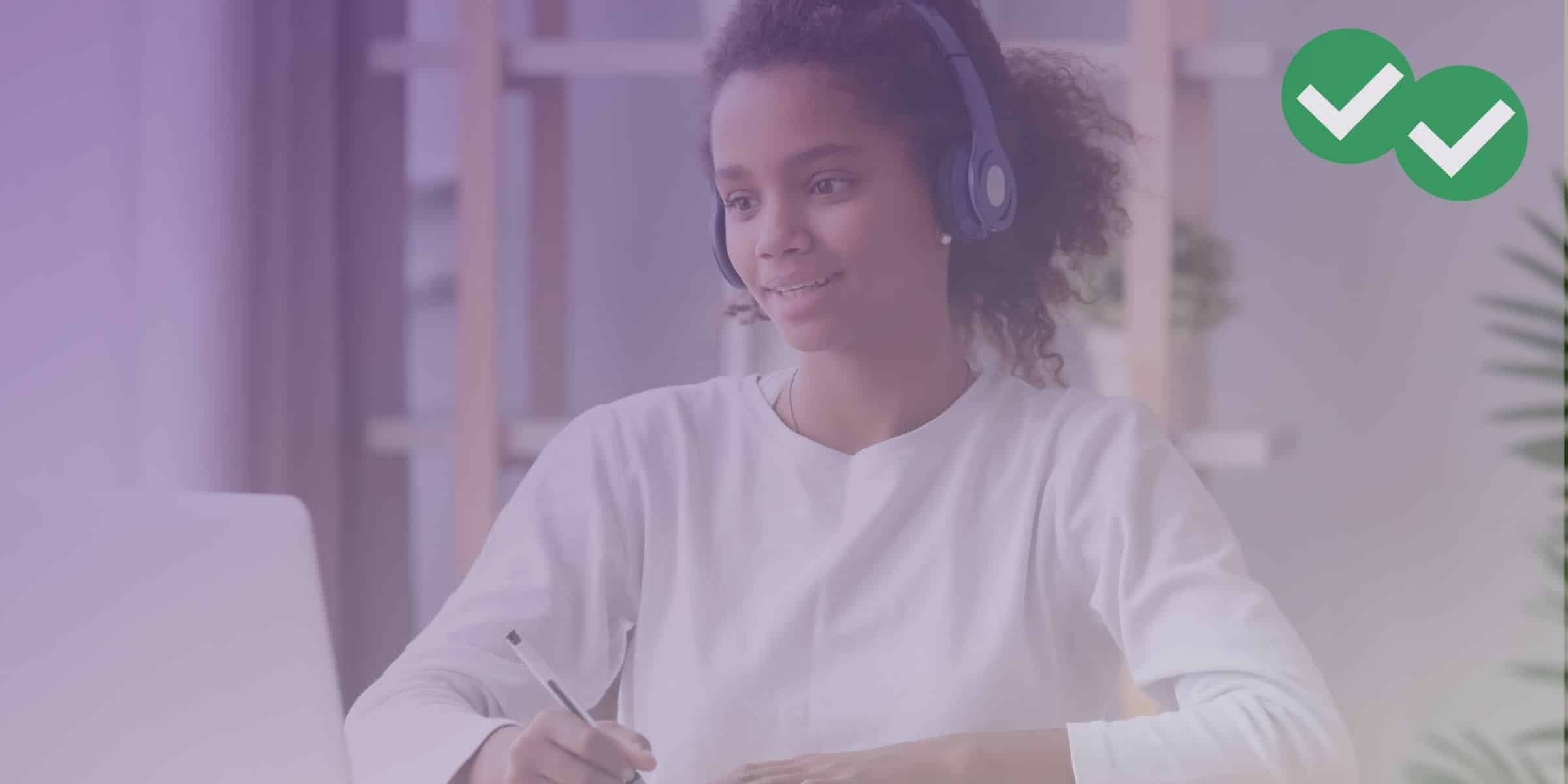 student smiling at laptop with headphones on while writing in notebook