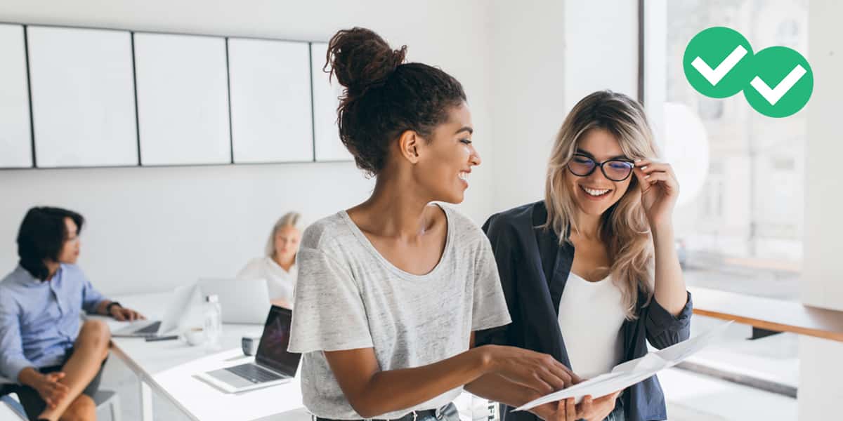 Young female executive discusses work with a colleague as their coworkers sit in the background in an office