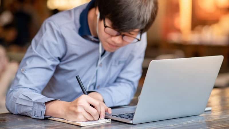 Man wearing earphones jots down notes on a notepad next to his laptop